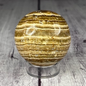 May include: A polished, banded, brown and white agate sphere on a clear acrylic stand. The sphere has a smooth, polished surface and is approximately 2 inches in diameter.