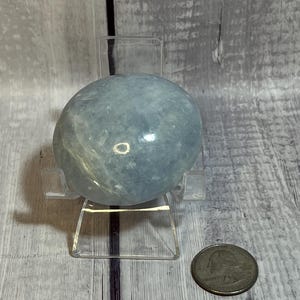 May include: A light blue, spherical crystal stone, possibly a gemstone, is displayed on a clear acrylic stand. A U.S. quarter coin is next to the stone for size comparison. The stone has a smooth, polished surface.