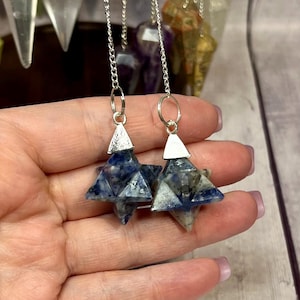 May include: Two blue and white sodalite pendulums with silver chains and triangular caps. The pendulums are star-shaped and held in a hand. Other pendulums are visible in the background.