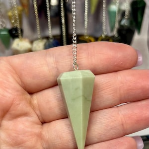 May include: A light green, faceted crystal pendulum suspended from a silver chain. The pendulum has a pointed, conical shape and is held in a hand. Other crystal pendulums are visible in the background.