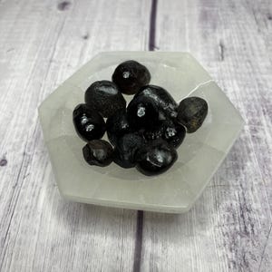 May include: A small white hexagonal dish filled with black obsidian tumbled stones. The stones are smooth and polished, and they have a dark, glassy appearance.
