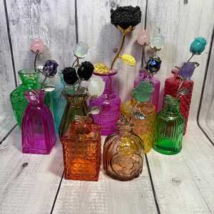 May include: A collection of colorful glass vases, each holding a decorative rose made of gemstones. The vases are in various shapes and colors, including pink, green, orange, and yellow. The roses are made of different gemstones, such as amethyst and rose quartz.