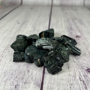 May include: A pile of black tourmaline crystals on a light wood surface. The crystals are rough and have a dark, almost black, color.