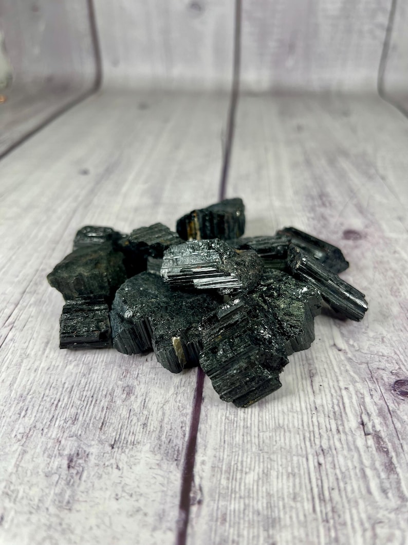 May include: A pile of black tourmaline crystals on a light wood surface. The crystals are rough and have a dark, almost black, color.