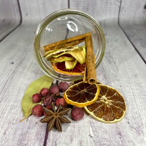 May include: A glass jar filled with dried fruit, cinnamon sticks, and star anise. The jar is tipped over, spilling the contents onto a wooden surface. The fruit includes orange slices, apple slices, and dried cranberries.