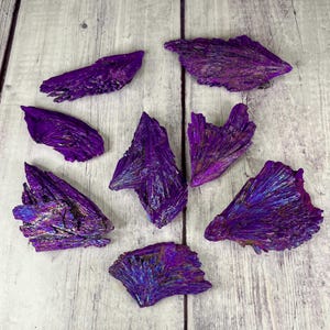 May include: A collection of eight purple iridescent kyanite crystals. The crystals are raw and have a natural, rough texture. They are arranged on a light wood surface.