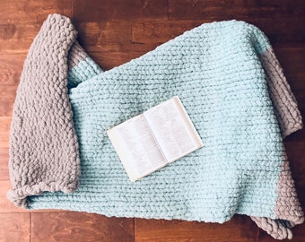 May include: A light blue and gray chunky knit throw blanket with a book on top of it.