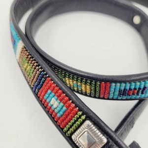 May include: A black leather belt with a colorful beaded design. The beads are arranged in a geometric pattern and include red, blue, green, and gold colors. A silver metal buckle is attached to the belt.