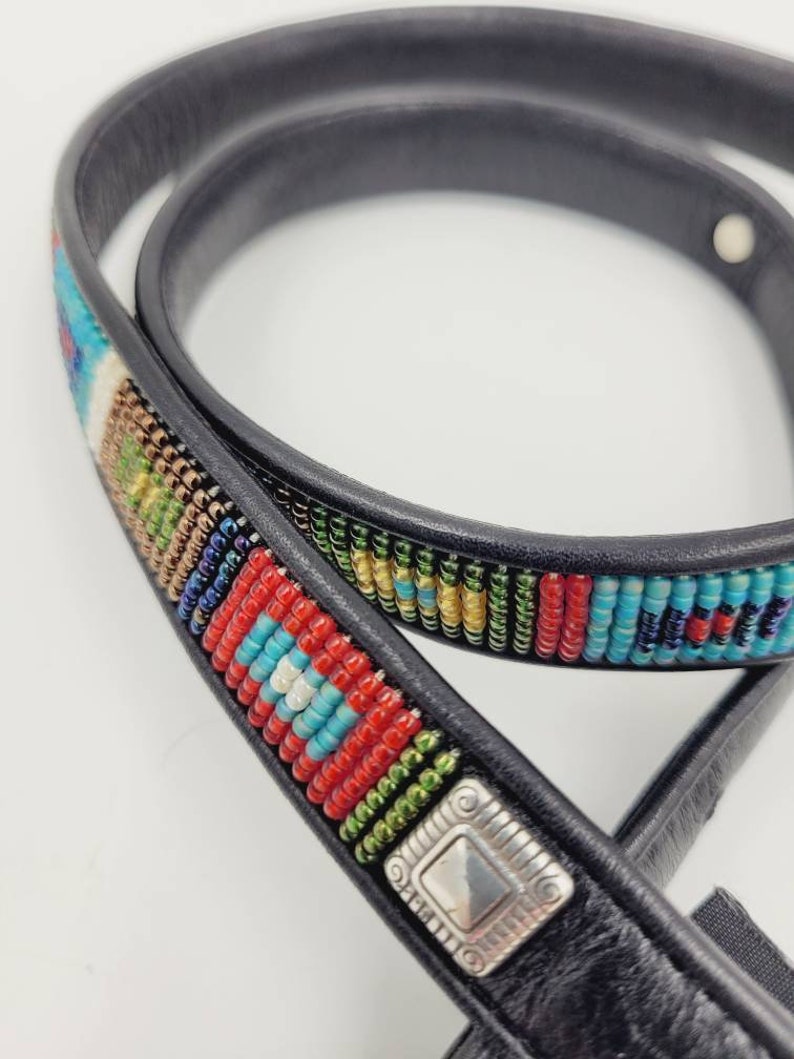 May include: A black leather belt with a colorful beaded design. The beads are arranged in a geometric pattern and include red, blue, green, and gold colors. A silver metal buckle is attached to the belt.