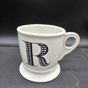 May include: White ceramic mug with a black and white patterned letter "R" on the front. The mug has a wide base and a handle.
