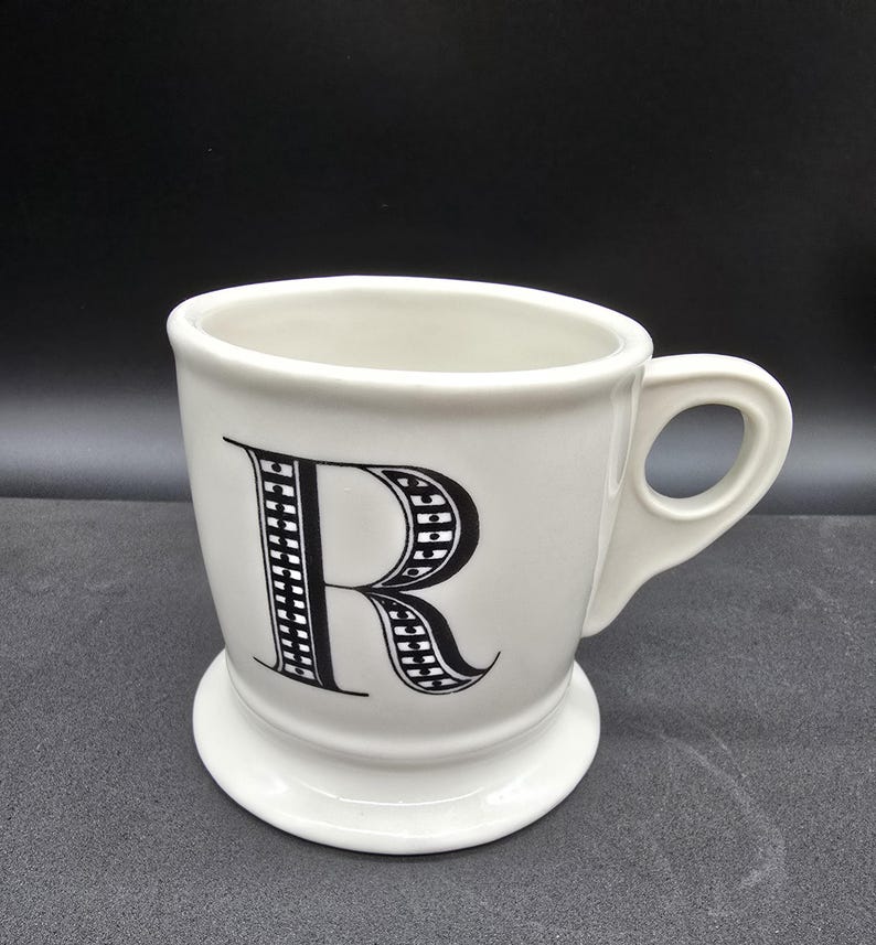 May include: White ceramic mug with a black and white patterned letter "R" on the front. The mug has a wide base and a handle.