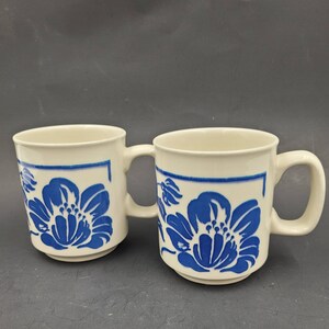 May include: Two white ceramic mugs with blue floral designs. The mugs have a square design with a blue border and a blue floral pattern inside the square. The mugs have a white background and blue handles.