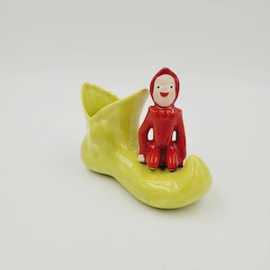 May include: A yellow ceramic shoe with a red elf figurine sitting on top. The elf is wearing a red hat and has a white face.