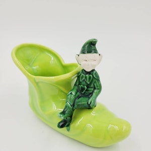 May include: A ceramic shoe-shaped planter in bright green, with a seated elf figurine. The elf is dressed in green with a white face. The planter is set against a white backdrop. The planter is a decorative item.