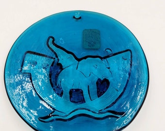 May include: A round, blue glass plate with an etched design of an elephant standing on a wave. The plate has a small, blue square sticker on the back.
