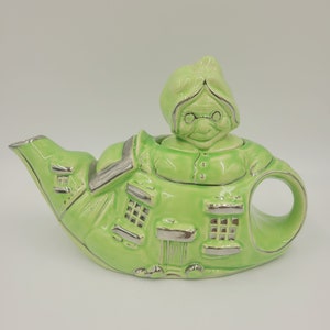 May include: A green ceramic teapot shaped like a woman with silver trim. The teapot has a lid with a woman's face on it. The teapot has a spout and a handle.