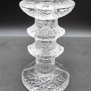 May include: A clear glass candle holder with a textured surface and a tiered design. The holder has a wide base and a narrow top.