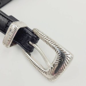 May include: A black leather belt with a silver buckle. The buckle has a decorative design with a textured pattern.