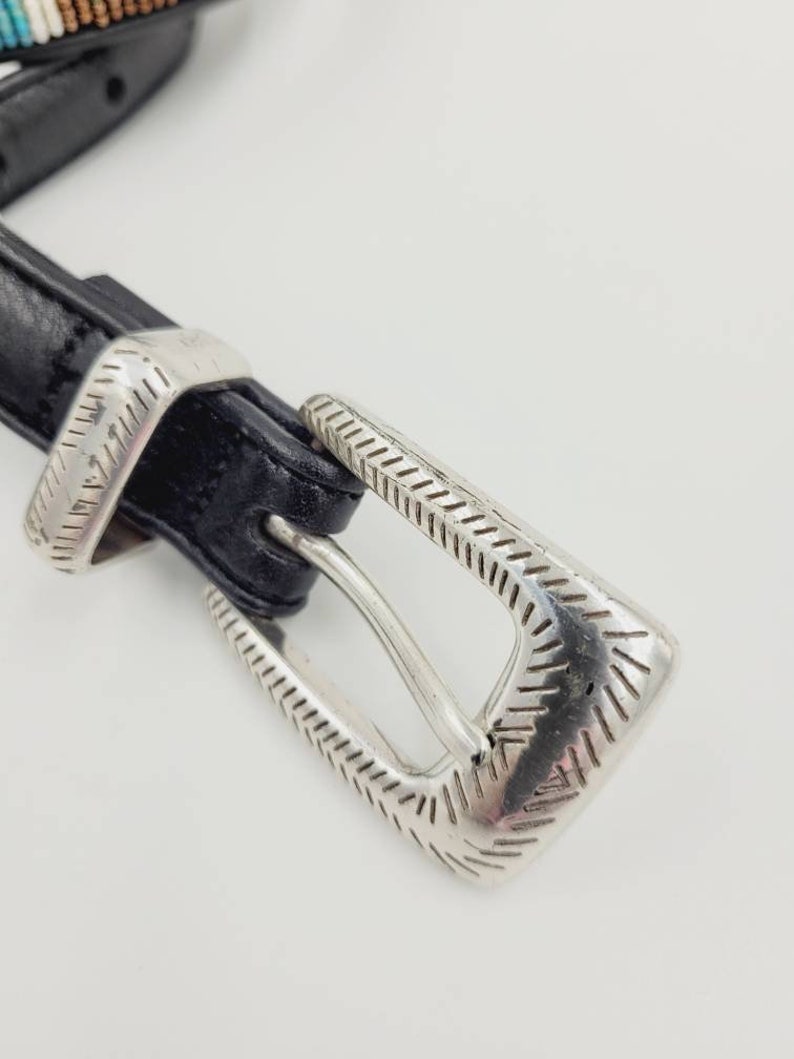 May include: A black leather belt with a silver buckle. The buckle has a decorative design with a textured pattern.