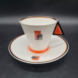 May include: A white teacup and saucer with a geometric design in black and orange. The cup has a triangular handle.