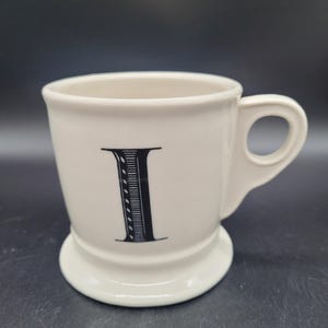 Puede incluir: Una taza de cer&aacute;mica blanca con una letra negra "I" impresa en la parte delantera. La taza tiene un asa redondeada y una base ligeramente acampanada.