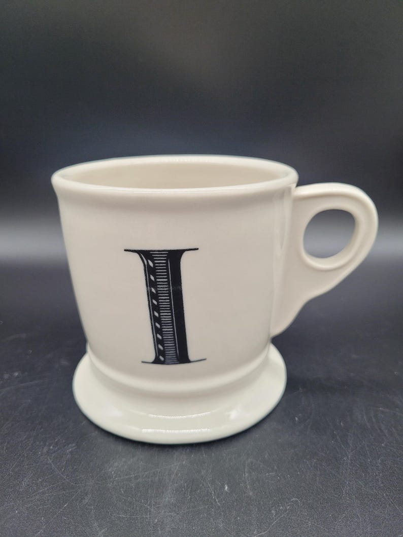 Puede incluir: Una taza de cer&aacute;mica blanca con una letra negra "I" impresa en la parte delantera. La taza tiene un asa redondeada y una base ligeramente acampanada.