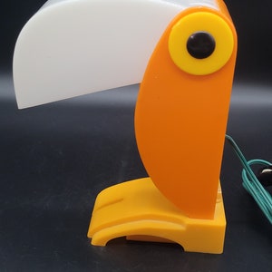 May include: Orange toucan-shaped desk lamp with a white shade and a yellow eye. The lamp is plugged in and has a green cord.