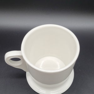 May include: A white ceramic mug with a wide base and a handle. The mug is empty and has a smooth, glossy finish.