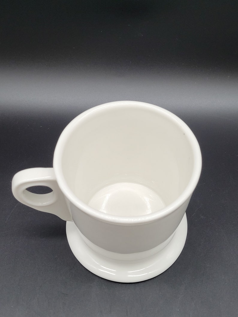 May include: A white ceramic mug with a wide base and a handle. The mug is empty and has a smooth, glossy finish.