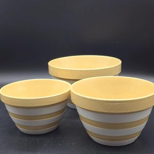 May include: Three ceramic mixing bowls in varying sizes. The bowls are yellow with white horizontal stripes. The rims are also yellow. The bowls are arranged on a dark surface.