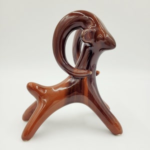 May include: A brown ceramic sculpture of a stylized ram with curved horns. The ram is standing on four legs and has a glossy finish.