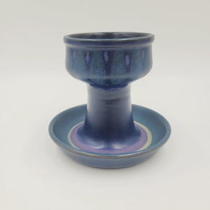May include: A blue ceramic candle holder with a wide base and a cup-shaped top. The base has concentric rings of purple and yellow. The holder is glazed with a glossy finish. The overall design is a unique and decorative piece.