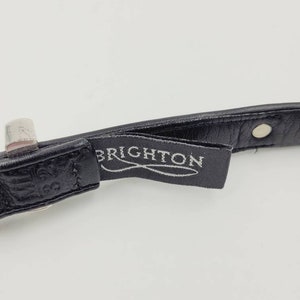 May include: Close-up of a black leather belt with a silver buckle and a black label that reads "BRIGHTON".