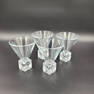 May include: Four clear glass martini glasses. Each glass features a conical bowl and a square, textured base. The glasses are displayed on a dark surface, highlighting their transparent design and elegant shape.