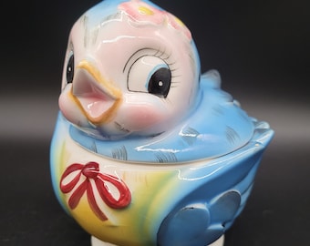 May include: A blue and yellow ceramic bird figurine with a pink flower on its head and a red bow around its neck. The bird has a white base and is a vintage style.