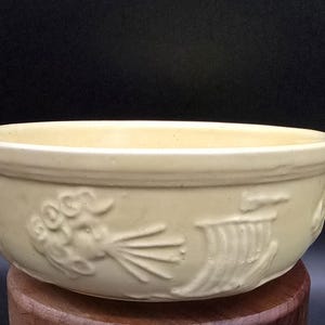 May include: A yellow ceramic bowl with a decorative design featuring a stylised floral motif. The bowl is sitting on a wooden surface.