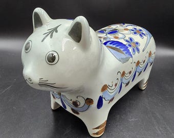May include: A ceramic cat figurine with a white base, featuring hand-painted blue and brown floral and scroll patterns across its back and sides. The cat has black painted eyes, whiskers, and ear details, with a smooth, glossy finish throughout.
