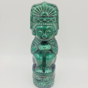 May include: A green ceramic statue of a seated figure with an elaborate headdress. The statue has a glossy finish and the word "Kancua" is inscribed on the base. The figure is in a seated position with hands clasped.