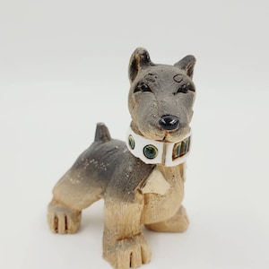 May include: A small ceramic dog figurine with a white collar and green gems. The dog is standing on all four paws and has a brown and grey colour scheme.