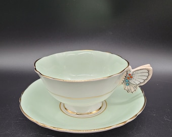 May include: A light green teacup and saucer set with a gold rim. The teacup has a butterfly-shaped handle with colorful accents.