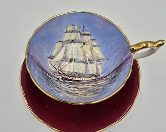 May include: A vintage teacup and saucer set featuring a blue and white illustration of a sailing ship on the cup. The saucer is a deep burgundy red with a gold rim.