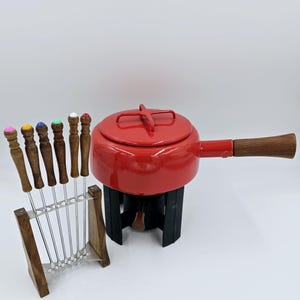 May include: Red enamel fondue pot with a wooden handle and a black metal stand. The pot has a lid with a cross-shaped handle. The set includes eight fondue forks with colorful plastic handles.