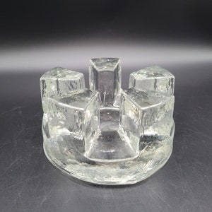 May include: A clear glass candle holder with a circular base and four square-shaped candle holders. The candle holders are arranged in a circle around the base.
