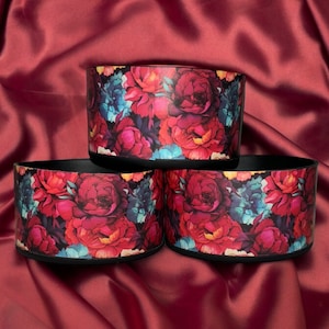 Puede incluir: Tres recipientes cilíndricos con un estampado floral de rosas rojas, rosas y azules. Los recipientes tienen una base e interior negros, y están dispuestos sobre una superficie de satén burdeos.