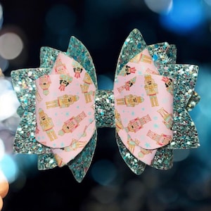 May include: A teal glitter hair bow with a pink and white fabric center featuring a repeating pattern of nutcracker soldiers.