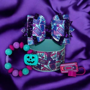 Peut inclure: Un ensemble d'accessoires sur le thème d'Halloween comprenant un nœud pour cheveux à paillettes violet et bleu sur le thème du cinéma d'horreur, un bracelet en silicone turquoise avec un charme en forme de citrouille et un porte-clés en silicone turquoise et rose avec un charme en forme de cassette VHS qui dit "HORROR".
