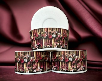 Puede incluir: Cuencos de cerámica blancos apilados y un platillo decorados con motivos florales. Los cuencos presentan un diseño de libros y flores en tonos marrón, rojo y rosa. El platillo tiene la inscripción "Cup Couture". El fondo es de tela burdeos oscuro.