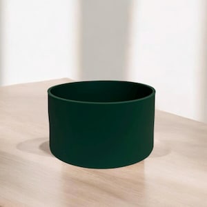 May include: A dark green cylindrical planter made of ceramic. The planter is sitting on a light wood surface.