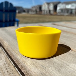 May include: A bright yellow silicone cup sits on a wooden table. The cup is empty and has a smooth, rounded surface.