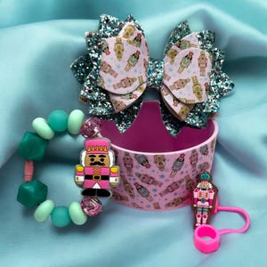 May include: A pink and teal nutcracker themed set includes a bracelet with teal and pink beads, a pink nutcracker charm, a pink cup with a nutcracker pattern, and a pink nutcracker keychain.